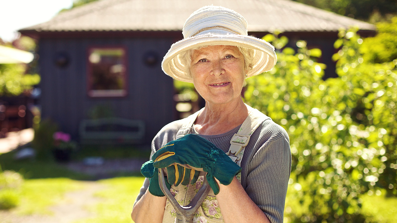 A woman standing in front of her house, holding a shovel with gardening gloves on, ready to tend to her garden with care.