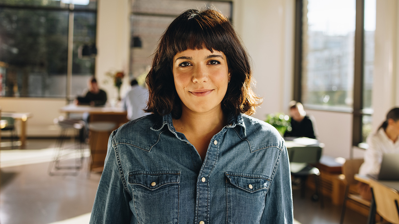 A 30-year-old brunette woman with a fringe, looking at the camera while standing in a professional workplace.