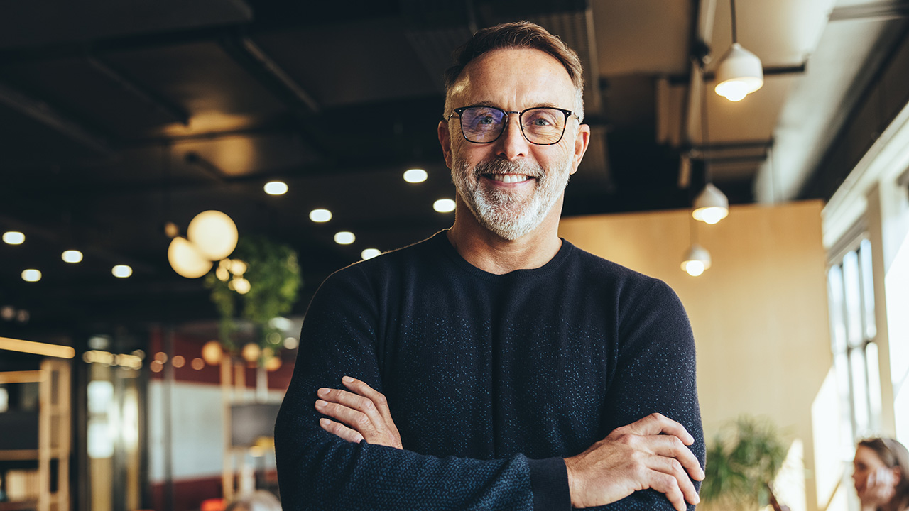 A 50-year-old male with glasses smiling at the camera while standing in his workplace.