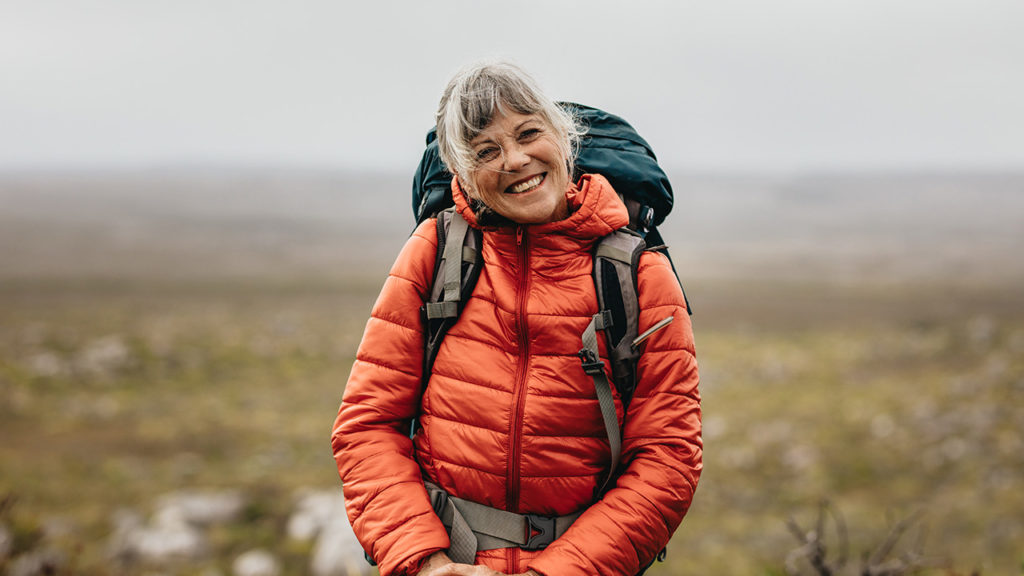 A smiling middle-aged woman wearing an orange puffer jacket and backpack, standing in front of a scenic view while hiking.