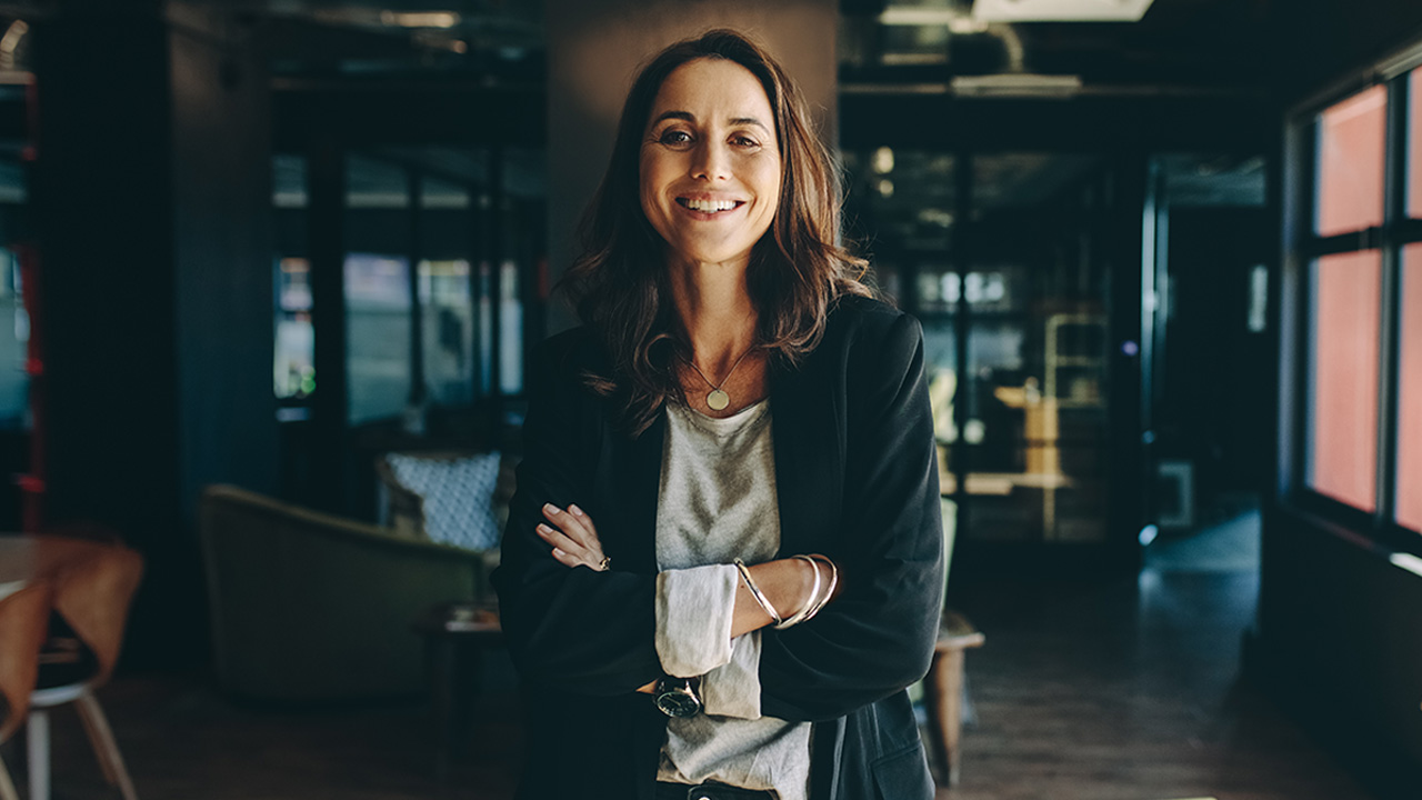 A 40-year-old female financial advisor standing confidently in a professional work environment, smiling with her arms crossed.