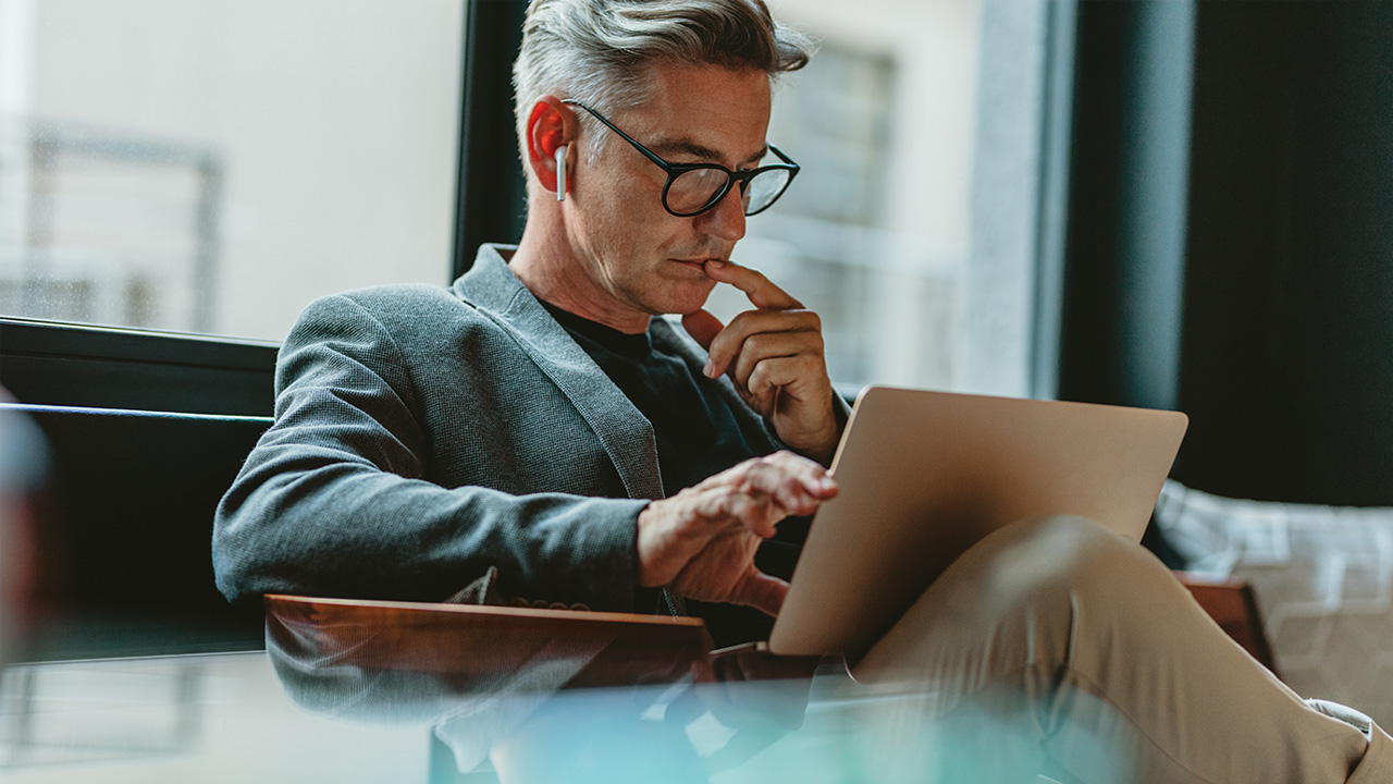 A 50-year-old male financial advisor with glasses and AirPods sitting, looking curiously at his laptop screen.