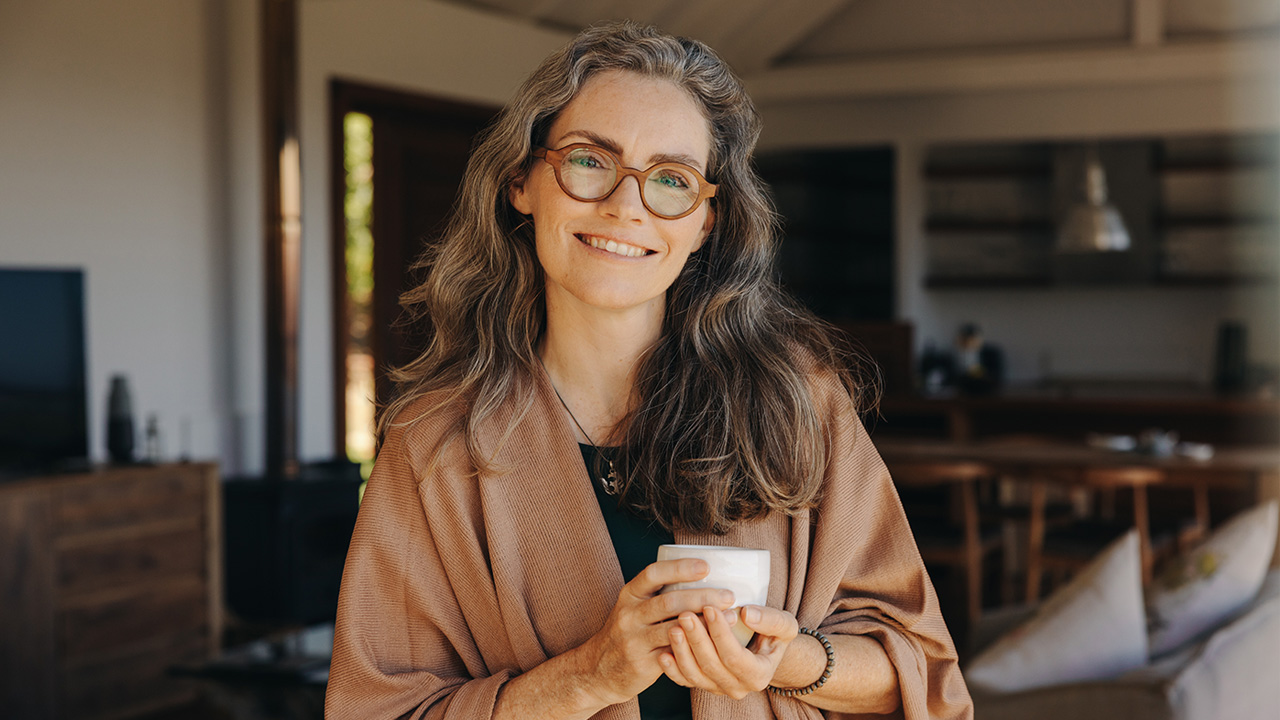 A middle-aged woman wearing glasses smiles warmly at the camera in the comfort of her cozy home