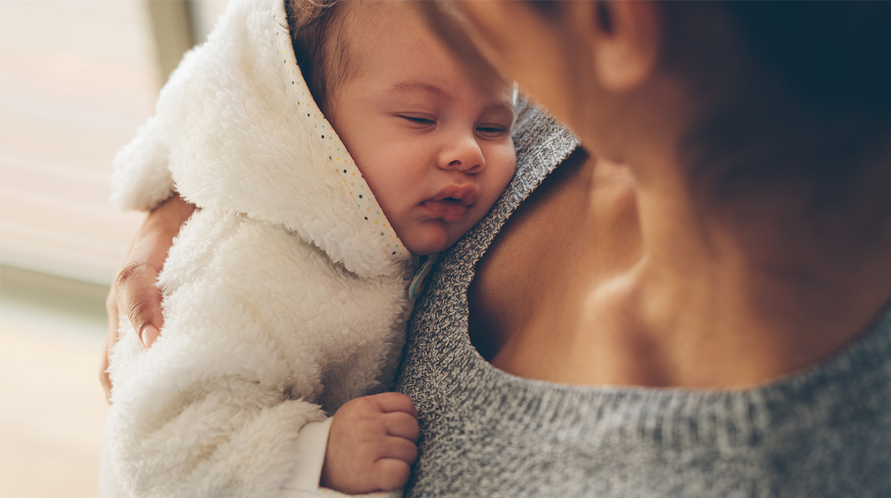 A close-up shot of a baby nestled in its mother's arms, wearing a white furry onesie.