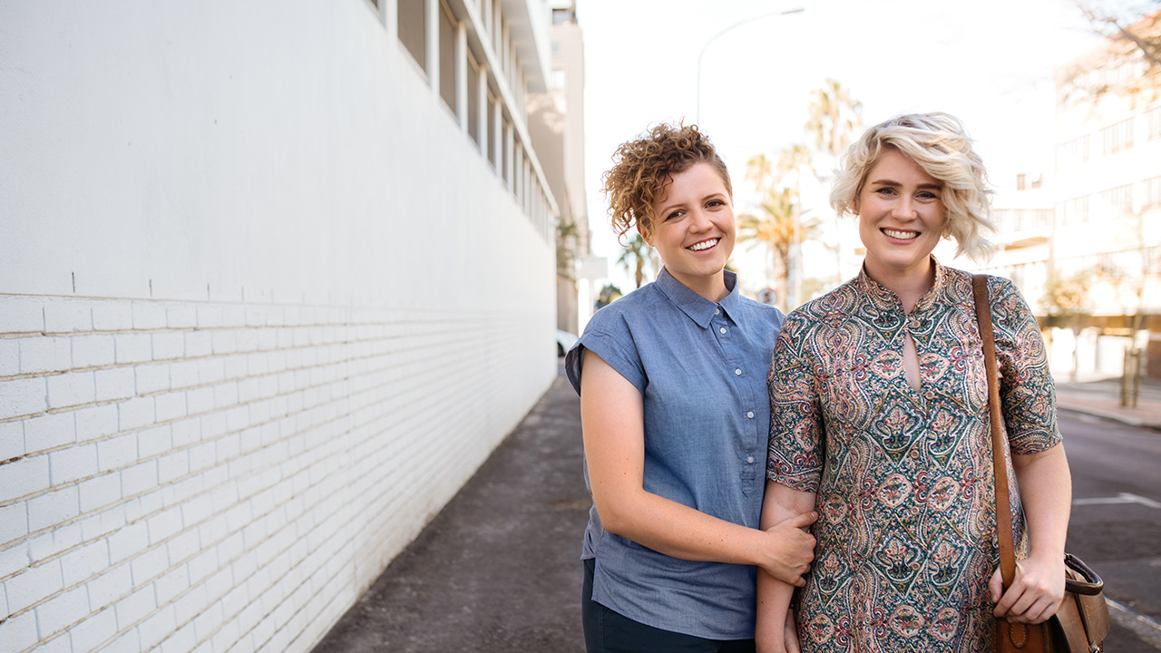 A same-sex female couple standing next to each other, holding hands on the street beside a white brick wall.