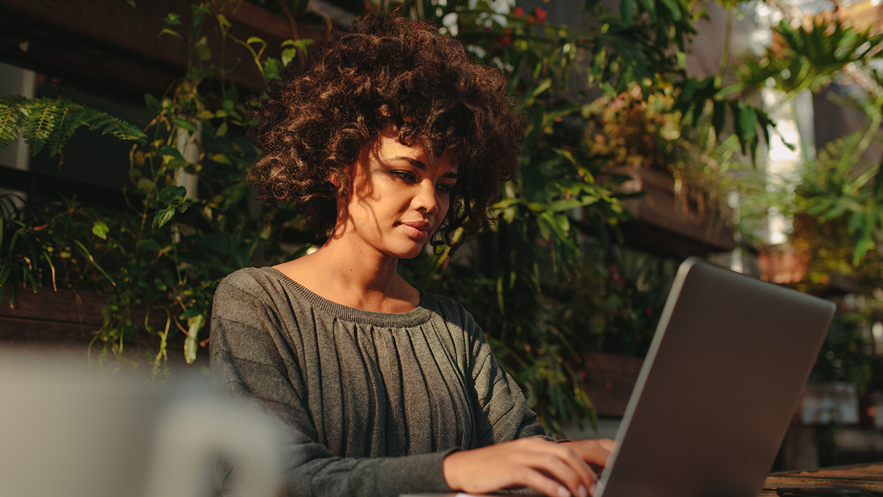 A woman sitting at a table outside, looking at her laptop with greenery behind her.