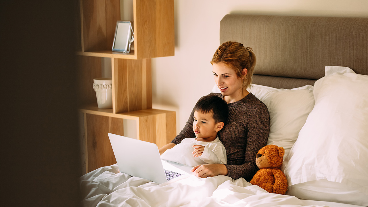 A mother and an Asian child sitting on a bed, looking at a laptop with a teddy bear beside them.