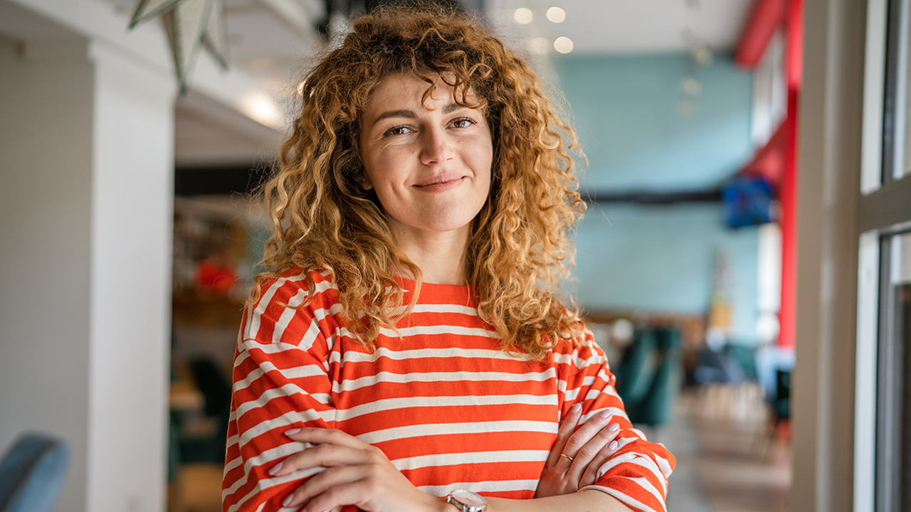 A woman with curly hair, wearing an orange striped top, with her arms crossed and smiling at the camera, with a blurred background.