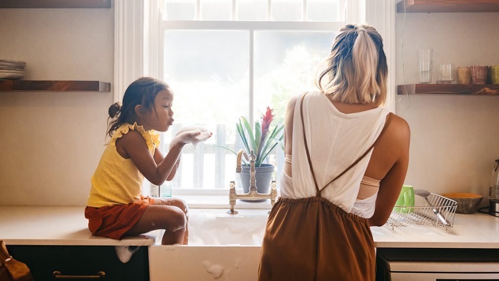 A woman and her daughter standing at the kitchen sink, with the daughter’s feet in the sink and bubbles blowing